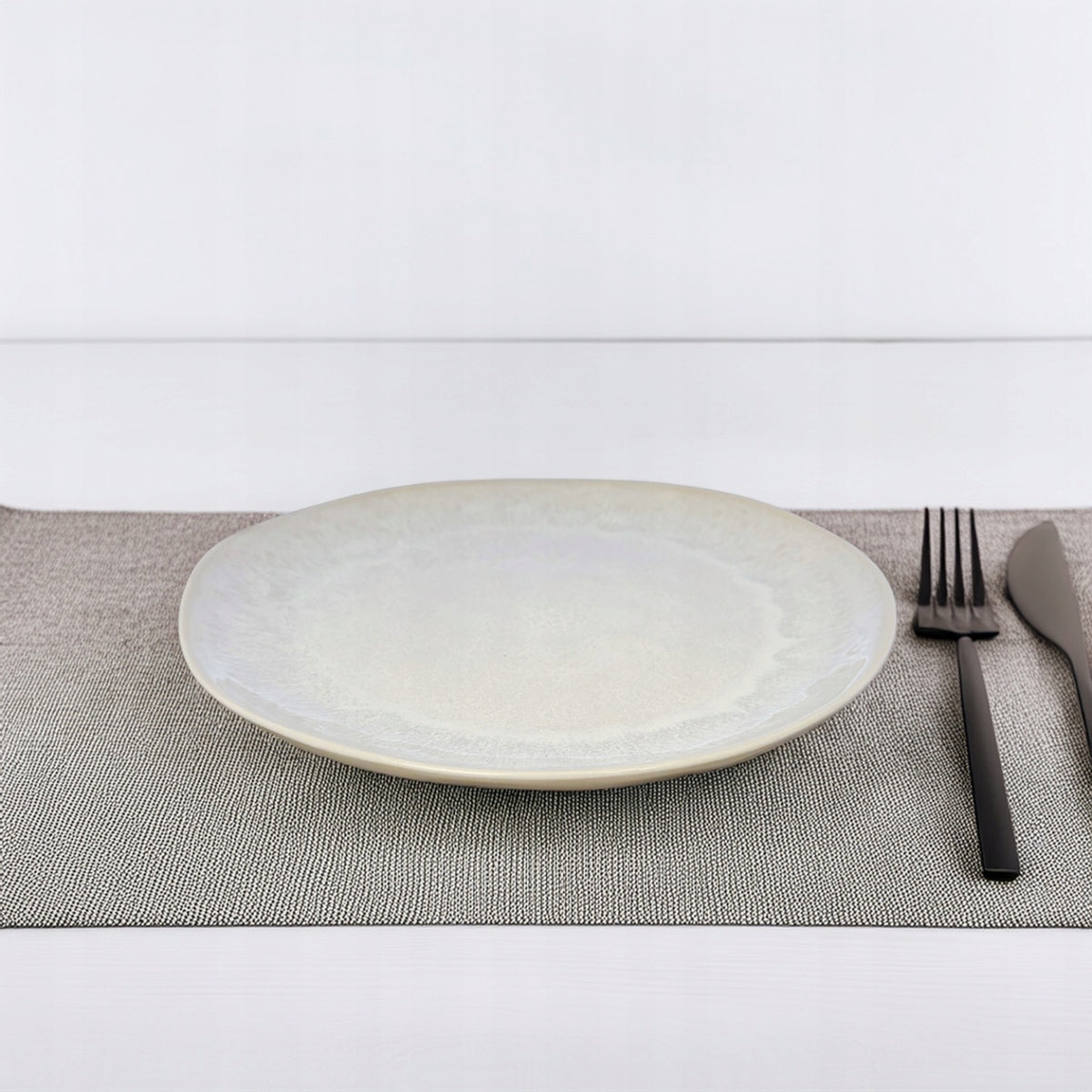 Minimalistyczna aranżacja stołu. Jasnokremowy talerz ceramiczny o subtelnie cieniowanej fakturze spoczywa na szarej, plecionej podkładce. Po prawej stronie naczynia ułożono eleganckie, matowe sztućce w czarnym kolorze (widelec i nóż). Kompozycja oparta jest na jasnym, czystym tle w nowoczesnym stylu.