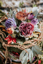 Jesienna kompozycja florystyczna (stroik) ułożona na bazie z mchu. W centrum znajdują się duże, sztuczne peonie w odcieniach głębokiego bordo i przygaszonego fioletu. Towarzyszą im mniejsze, rude pąki, czerwone jesienne liście (klonu), szarozielone listki oraz długie, opadające pędy koralowego szarłatu (amaranthusa)