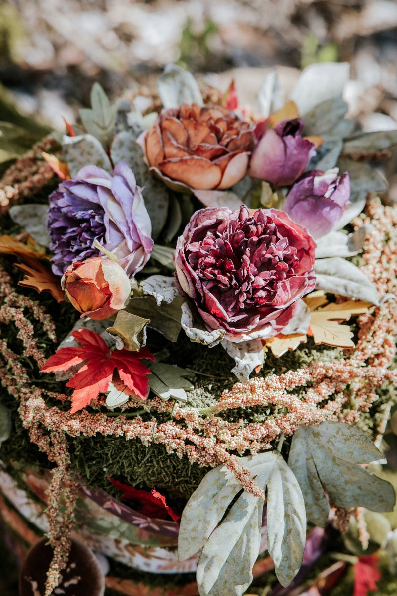 Jesienna kompozycja florystyczna (stroik) ułożona na bazie z mchu. W centrum znajdują się duże, sztuczne peonie w odcieniach głębokiego bordo i przygaszonego fioletu. Towarzyszą im mniejsze, rude pąki, czerwone jesienne liście (klonu), szarozielone listki oraz długie, opadające pędy koralowego szarłatu (amaranthusa)