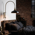 Lampa ścienna w stylu industrialnym (kinkiet) zamontowana nad łóżkiem w sypialni. Lampa ma wygięte, mosiężne ramię i czarny, metalowy klosz. Wisi nad drewnianym wezgłowiem łóżka zasłanego pościelą z ciemnego, gniecionego lnu. W tle znajduje się surowa ściana z czerwonej cegły oraz okno ze szprosami.