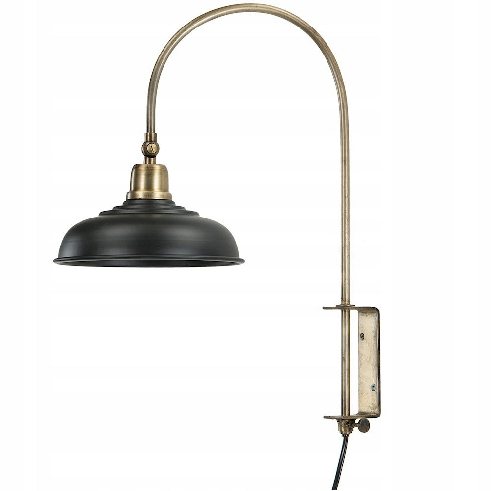 Zdjęcie produktowe na białym tle (packshot). Lampa ścienna w stylu loft/industrialnym. Składa się z długiego, łukowato wygiętego ramienia w kolorze patynowanego mosiądzu oraz czarnego, półokrągłego, metalowego klosza. Konstrukcja pozwala na montaż bezpośrednio do ściany za pomocą płaskiej bazy, z której wychodzi czarny przewód zasilający.