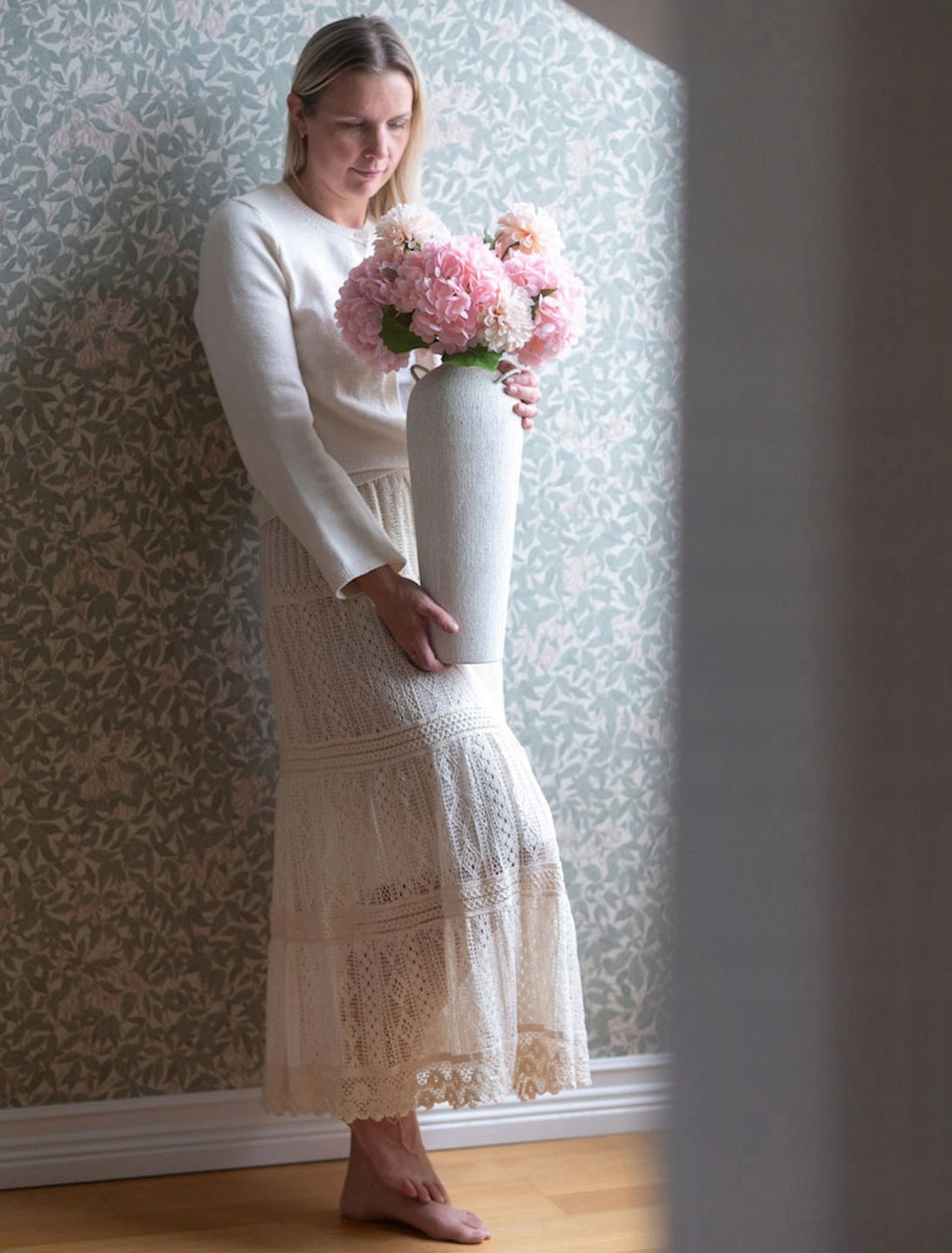 Bosa kobieta w kremowym swetrze i długiej, jasnej, koronkowej spódnicy w stylu boho stoi oparta o ścianę wykończoną kwiecistą, zieloną tapetą. Trzyma w dłoniach na wysokości bioder wysoki, fakturowany wazon ceramiczny, w którym znajduje się gęsty bukiet sztucznych, różowych kwiatów. Zdjęcie ma ciepły, romantyczny klimat.