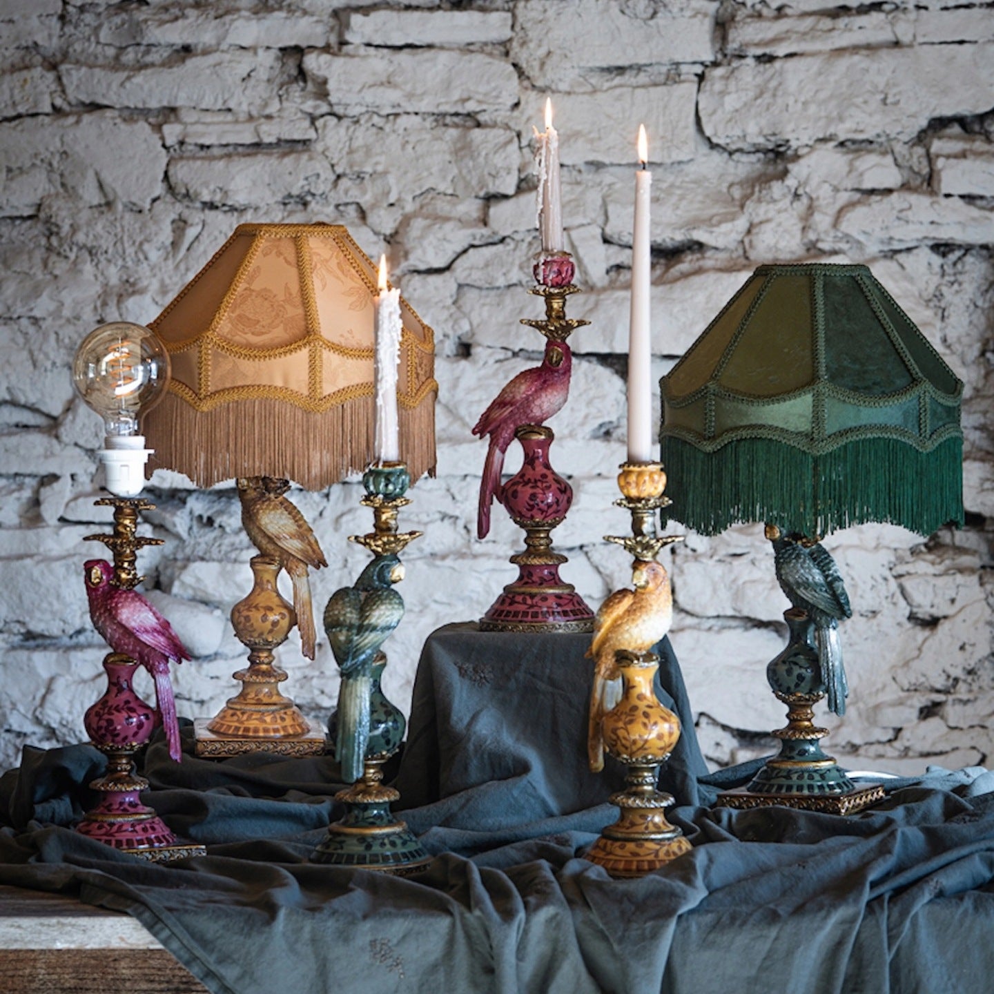 Nastrojowa, artystyczna kompozycja na tle surowego, białego kamiennego muru. Na stole przykrytym drapowanym, ciemnoszarym lnem wyeksponowano kolekcję dekoracji w stylu vintage. Są to lampy i świeczniki o podstawach z figurkami papug w różnych kolorach (zielonym, musztardowym, amarantowym). Dwie lampy mają eleganckie abażury z frędzlami (miodowy i ciemnozielony), jedna ma wkręconą ozdobną, odsłoniętą żarówkę, a w dwóch wysokich świecznikach płoną jasne, klasyczne świece.