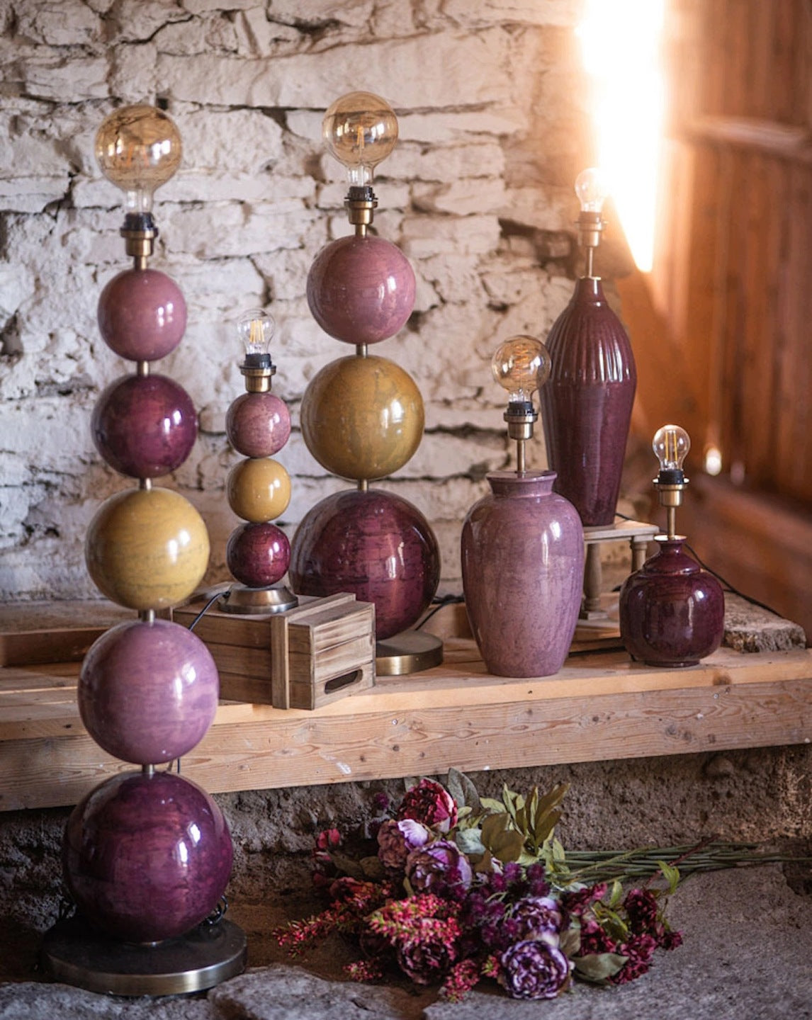 Surowa, rustykalna aranżacja na tle starego, pobielanego muru z kamienia. Na drewnianej półce i małych skrzynkach wyeksponowano bogatą kolekcję różnorodnych, ceramicznych podstaw do lamp w odcieniach purpury, zgaszonego różu oraz musztardowej żółci. Podstawy przyjmują różne formy – od smukłych butli, przez pękate wazy, aż po oryginalne konstrukcje z ułożonych na sobie kul. W oprawkach zamontowane są ozdobne, odsłonięte żarówki typu Edison z widocznymi żarnikami, a na pierwszym planie leży bukiet sztucznych,