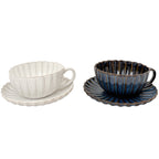 Zdjęcie produktowe typu packshot wykonane na jednolitym, białym tle. Przedstawia zestaw dwóch ceramicznych filiżanek z dopasowanymi spodkami. Naczynia charakteryzują się elegancką, prążkowaną fakturą oraz nieregularnymi, pofalowanymi brzegami przypominającymi płatki kwiatów. Lewa filiżanka jest w kolorze złamanej bieli, natomiast prawa pokryta jest lśniącym, głębokim, granatowym szkliwem z brązowymi akcentami na rantach.