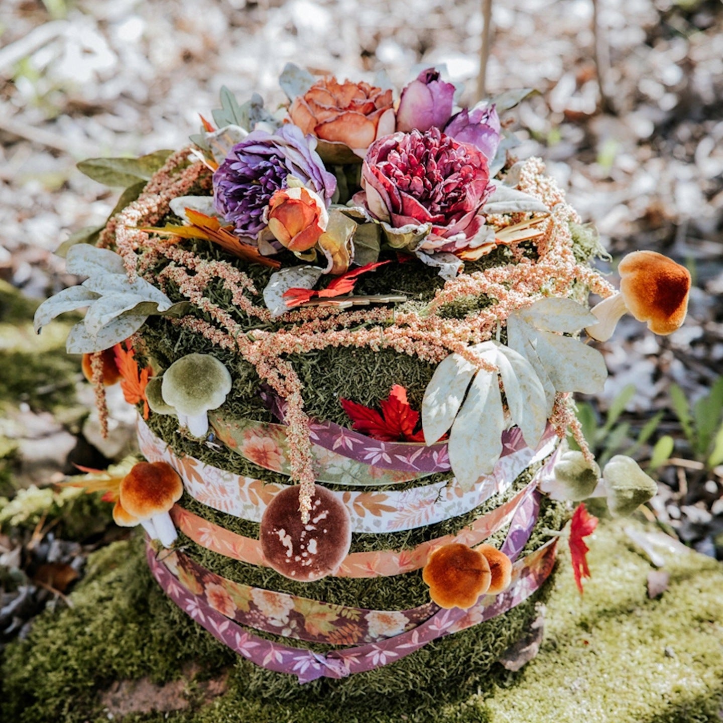 Niezwykła kompozycja florystyczna ułożona w leśnym otoczeniu. Ma kształt wysokiego walca (przypominającego tort) pokrytego zielonym mchem. Boki "tortu" są owinięte kilkoma warstwami ozdobnych tasiemek w jesienne, roślinne wzory. Z mchu wystają małe, dekoracyjne grzybki z welurowymi kapeluszami w kolorze zielonym i brązowym oraz sztuczne jesienne liście. Na wierzchu znajduje się bukiet purpurowych i bordowych kwiatów, z którego zwisa długi, koralowy szarłat.