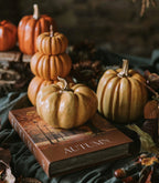 Zbliżenie na przytulną, jesienną dekorację. Na pierwszym planie, na książce z napisem "AUTUMN", leży pękata, błyszcząca, żółta dynia ze złotym ogonkiem. W tle widać kolejną żółtą dynię oraz wieżę z dyń w kolorze pomarańczowym. Aranżacja jest dopełniona suchymi, jesiennymi liśćmi.