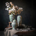Nastrojowe, artystyczne zdjęcie typu martwa natura (chiaroscuro). Na okrągłym, drewnianym stole przykrytym pogniecionym, szaro-zielonym lnem stoją trzy zielone naczynia ceramiczne w białe, roślinne wzory: dwa wysokie wazony i jedna pękata waza z pokrywką. Z najwyższego wazonu, stojącego na małym, drewnianym podeście, wylewa się bogaty bukiet sztucznych lub suszonych kwiatów – piwonii i drobnych, białych kwiatuszków. Tło jest ciemne, niemal czarne.