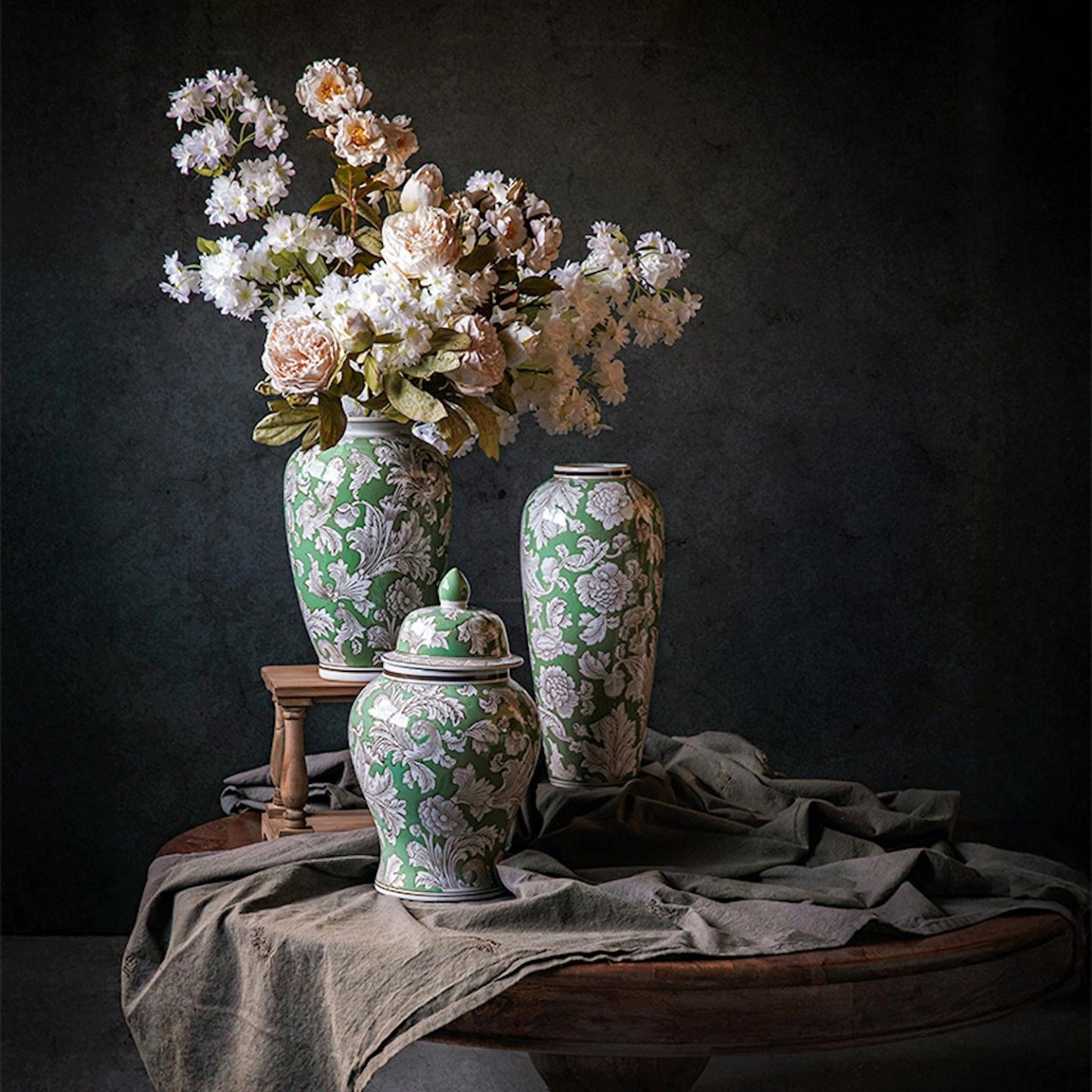 Nastrojowe, artystyczne zdjęcie typu martwa natura (chiaroscuro). Na okrągłym, drewnianym stole przykrytym pogniecionym, szaro-zielonym lnem stoją trzy zielone naczynia ceramiczne w białe, roślinne wzory: dwa wysokie wazony i jedna pękata waza z pokrywką. Z najwyższego wazonu, stojącego na małym, drewnianym podeście, wylewa się bogaty bukiet sztucznych lub suszonych kwiatów – piwonii i drobnych, białych kwiatuszków. Tło jest ciemne, niemal czarne.