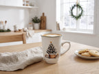 Jasne, przytulne wnętrze kuchni z drewnianym stołem na pierwszym planie. Na stole stoi kubek z kawą cappuccino z delikatnym latte art w kształcie serca, ozdobiony świątecznym motywem choinki w koszu. Obok leży lniana serweta oraz talerzyk z kruchymi ciasteczkami. W tle widać jasną kuchnię i wieniec świerkowy powieszony w oknie