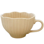 Zdjęcie produktowe na białym tle (packshot). Pojemny kubek (lub duża filiżanka) ceramiczny w jasnobeżowym (kremowym) kolorze. Naczynie charakteryzuje się pękatym, zaokrąglonym kształtem, ryflowanymi ściankami zewnętrznymi, falistą górną krawędzią oraz gładkim, wygodnym uchem.