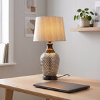 Jasne, nowoczesne domowe biuro (home office). Na prostym, dębowym biurku stoi elegancka lampa stołowa z zapalonym, jasnobeżowym abażurem. Jej podstawa jest wykonana ze srebrnego, strukturalnego szkła. Przed lampą, na blacie, leży zamknięty, ciemnoszary laptop. W tle znajduje się jasnoszara ściana z dwiema minimalistycznymi grafikami oraz drewniana półka, na której stoi mała roślina doniczkowa (pieniążek).