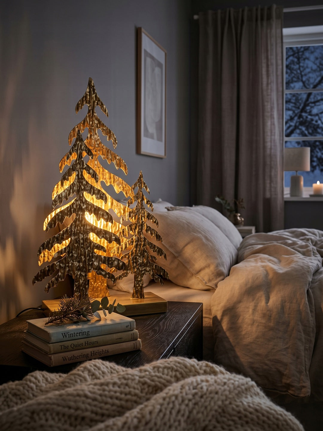 Niezwykle przytulna, wieczorna aranżacja sypialni w stylu hygge. Złota lampa w kształcie choinek z młotkowanego metalu stoi na ciemnej szafce nocnej, dając bardzo ciepłe, relaksujące światło. Obok leżą stosiki książek (w tym "Wuthering Heights") z małymi gałązkami na wierzchu. W tle widoczne jest zasłane, zachęcające łóżko z pościelą z naturalnego lnu oraz rzuconym grubym, wełnianym kocem.