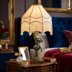 Luksusowa sypialnia w stylu glamour. Na eleganckiej, drewnianej szafce nocnej świeci nastrojowa lampa stołowa z podstawą w kształcie zielonej papugi i jasnym abażurem wykończonym długimi frędzlami z chwostami. Obok lampy stoi małe lusterko w złotej ramce oraz kryształowa buteleczka perfum z pompką. W głębi znajduje się duże łóżko z welurowym, granatowym zagłówkiem pikowanym i bordową, welurową narzutą.