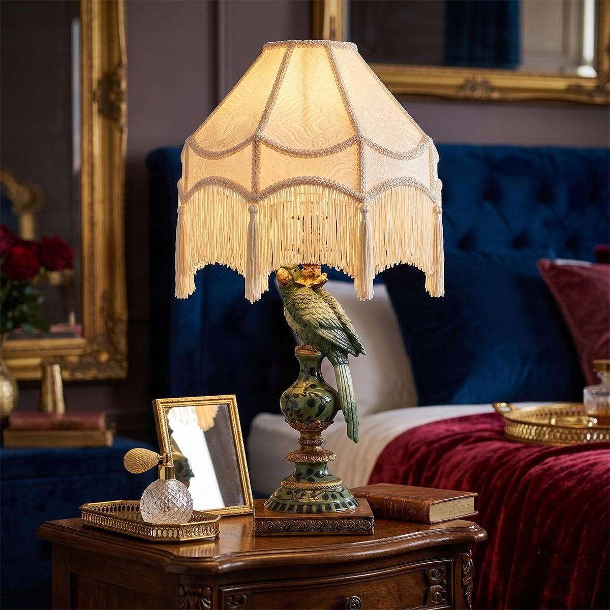 Luksusowa sypialnia w stylu glamour. Na eleganckiej, drewnianej szafce nocnej świeci nastrojowa lampa stołowa z podstawą w kształcie zielonej papugi i jasnym abażurem wykończonym długimi frędzlami z chwostami. Obok lampy stoi małe lusterko w złotej ramce oraz kryształowa buteleczka perfum z pompką. W głębi znajduje się duże łóżko z welurowym, granatowym zagłówkiem pikowanym i bordową, welurową narzutą.