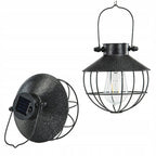 Zdjęcie produktowe na białym, czystym tle (packshot). Zestawienie dwóch rzutów na wiszącą lampę solarną w stylu loftowym. Lampa z prawej strony wisi prosto na metalowym uchwycie, ukazując przypominający dzwonek kloszyk, drucianą osłonę oraz stylizowaną żarówkę LED. Lampa z lewej strony jest pochylona, aby wyraźnie wyeksponować górną część klosza, gdzie zamontowany jest niewielki, kwadratowy panel słoneczny oraz przycisk zasilania. 