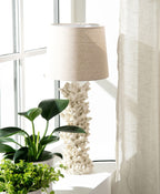 Lampa stołowa z podstawą w kształcie białego koralowca, zwieńczona jasnobeżowym, materiałowym abażurem. Lampa stoi na białym blacie lub parapecie w pełnym słońcu, tuż przy oknie z lnianą zasłoną. Obok niej ustawiono zielone rośliny doniczkowe. Jasna, świeża aranżacja wprowadzająca do wnętrza letni klimat