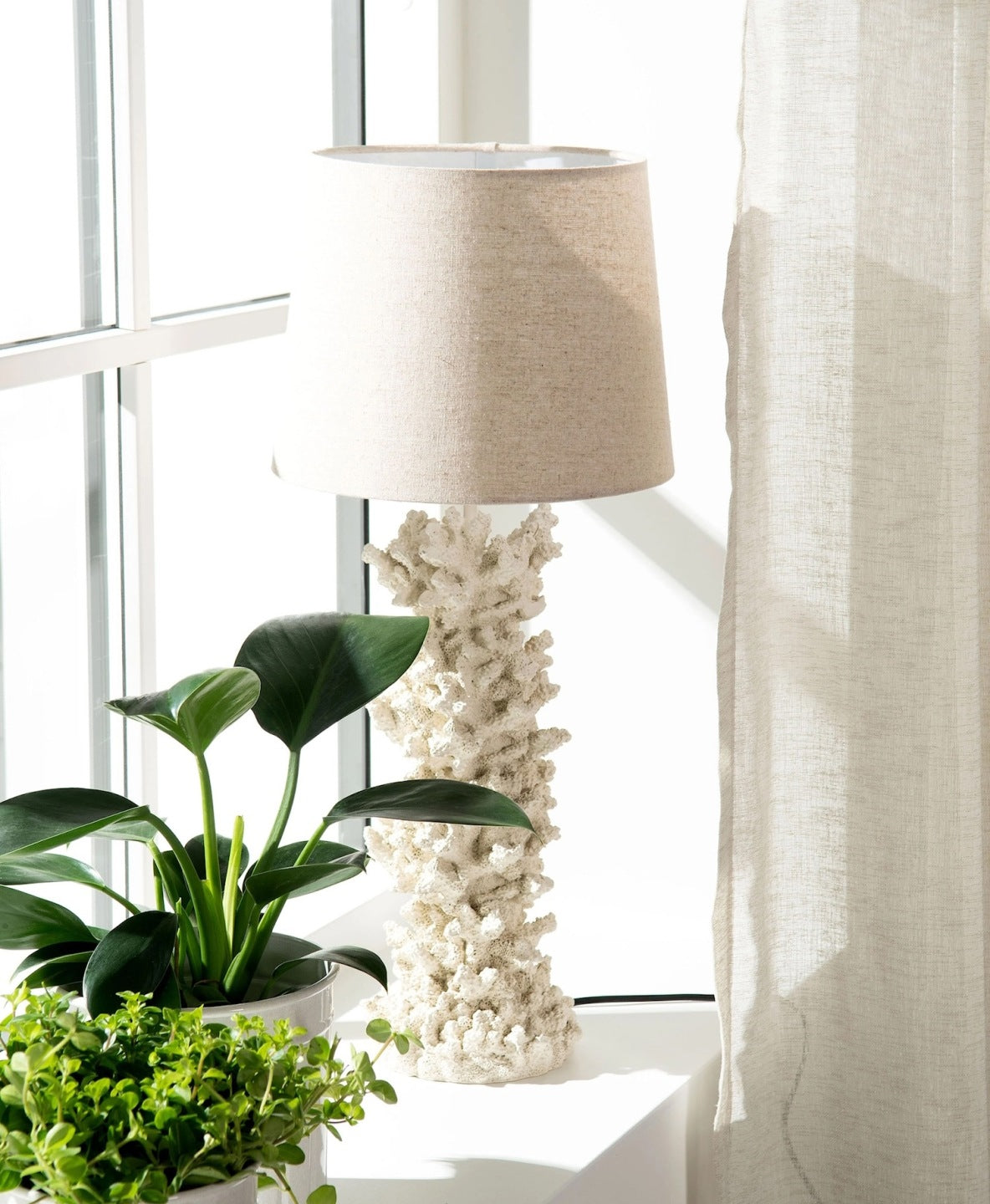 Lampa stołowa z podstawą w kształcie białego koralowca, zwieńczona jasnobeżowym, materiałowym abażurem. Lampa stoi na białym blacie lub parapecie w pełnym słońcu, tuż przy oknie z lnianą zasłoną. Obok niej ustawiono zielone rośliny doniczkowe. Jasna, świeża aranżacja wprowadzająca do wnętrza letni klimat