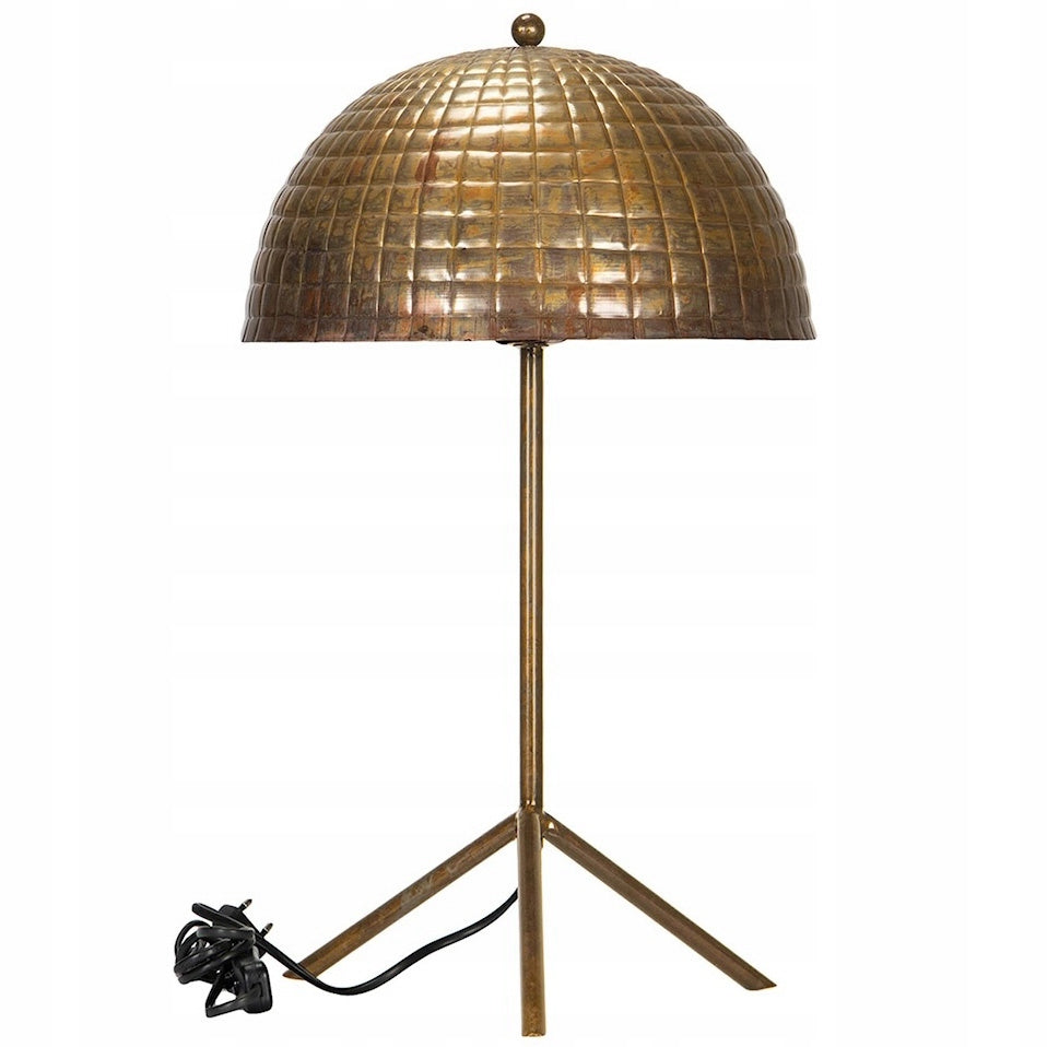 Zdjęcie produktowe na białym, czystym tle (packshot). Lampa stołowa wykonana w całości z postarzanego metalu (imitacja mosiądzu lub miedzi) o surowym, loftowym charakterze. Lampa opiera się na prostym trójnogu połączonym z pionową nóżką. Półokrągły klosz charakteryzuje się wyrazistą, tłoczoną fakturą w drobną kostkę/kratkę, z widocznymi, rdzawymi przebarwieniami. Widoczny jest czarny kabel zasilający z wtyczką.