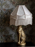 Lampa stołowa z jasnokremowym, klasycznym abażurem ozdobionym długimi frędzlami. Podstawa lampy ma kształt rzeźby siedzącego lamparta (geparda) w kolorze antycznego złota z ciemnymi cętkami. Lampa stoi na ciemnym blacie, a tło stanowi tapeta w gęsty, roślinno-kwiatowy wzór utrzymany w stonowanej kolorystyce