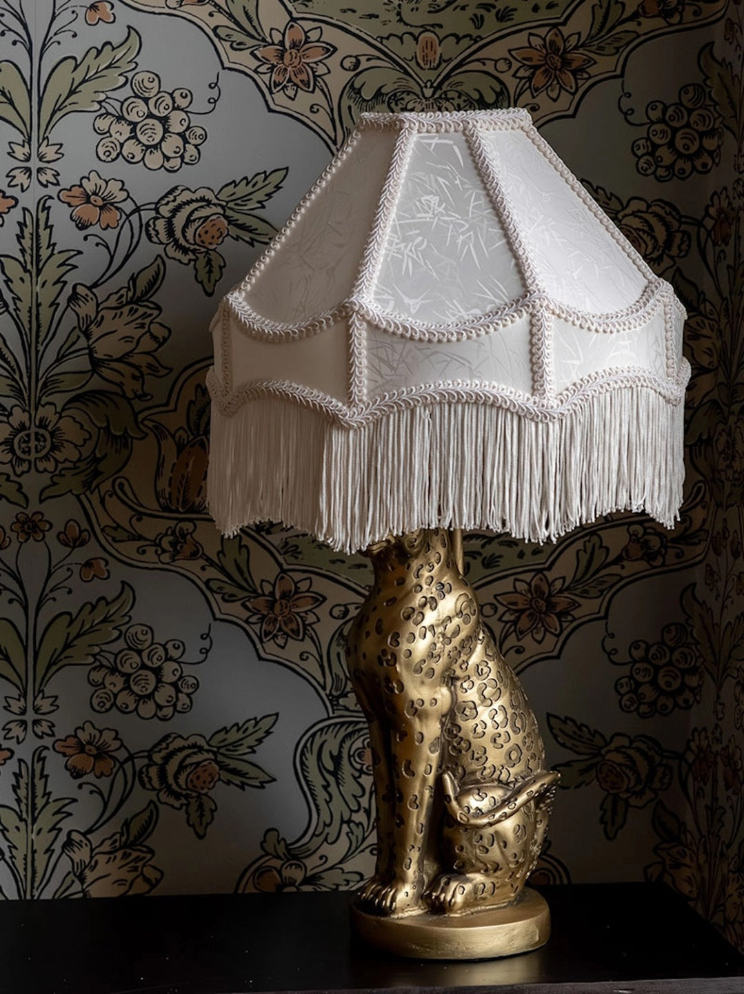 Lampa stołowa z jasnokremowym, klasycznym abażurem ozdobionym długimi frędzlami. Podstawa lampy ma kształt rzeźby siedzącego lamparta (geparda) w kolorze antycznego złota z ciemnymi cętkami. Lampa stoi na ciemnym blacie, a tło stanowi tapeta w gęsty, roślinno-kwiatowy wzór utrzymany w stonowanej kolorystyce