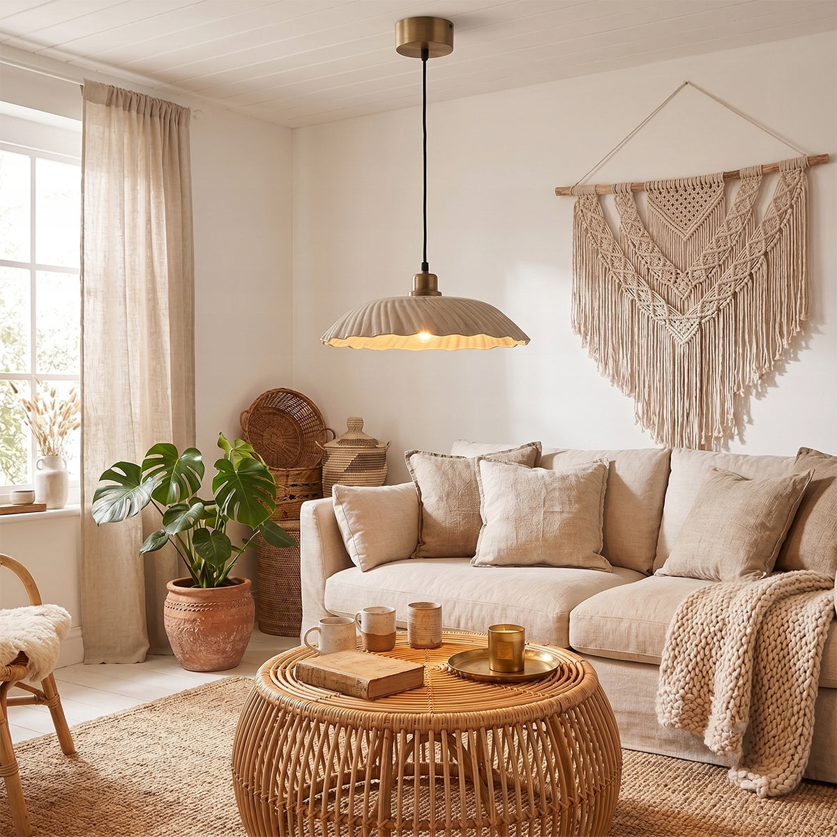 Przytulny salon urządzony w stylu boho. Centralnym punktem oświetlenia jest wisząca lampa z beżowym, karbowanym kloszem z ceramiki i mosiężną podsufitką. Lampa wisi nad okrągłym, rattanowym stolikiem kawowym. W tle znajduje się jasna, beżowa kanapa z lnianymi poduszkami, a na białej ścianie wisi duża, ręcznie pleciona makrama. Całość kompozycji dopełnia duża roślina doniczkowa – monstera w glinianej donicy.