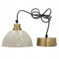 Zdjęcie produktowe na białym, gładkim tle (packshot). Lampa wisząca w stylu retro. Posiada błyszczący, jasnokremowy klosz o wyraźnie falistej (karbowanej) fakturze. Klosz zamocowany jest w szczotkowanej, mosiężnej oprawce, od której odchodzi zwinięty w pętle czarny przewód zasilający. Przewód zakończony jest okrągłą, metalową podsufitką w kolorze mosiądzu.