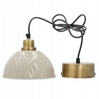 Zdjęcie produktowe na białym, gładkim tle (packshot). Lampa wisząca w stylu retro. Posiada błyszczący, jasnokremowy klosz o wyraźnie falistej (karbowanej) fakturze. Klosz zamocowany jest w szczotkowanej, mosiężnej oprawce, od której odchodzi zwinięty w pętle czarny przewód zasilający. Przewód zakończony jest okrągłą, metalową podsufitką w kolorze mosiądzu.
