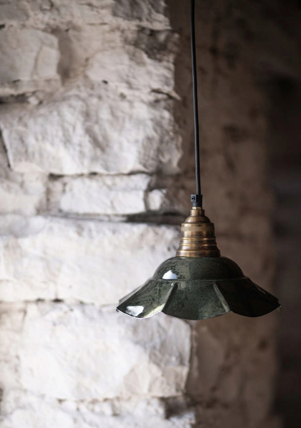 Klimatyczne ujęcie lampy wiszącej w rustykalnym wnętrzu. Lampa z lśniącym, zielonym, pofalowanym kloszem i mosiężną oprawą wisi na czarnym kablu. Tło stanowi surowa, nierównomierna ściana zbudowana z jasnego, naturalnego kamienia, co idealnie kontrastuje z eleganckim i starodawnym wyglądem oprawy oświetleniowej. Z błyszczącej powierzchni klosza odbija się światło.