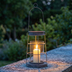 Zapalony lampion druciany stojący na kamiennym murku o zmierzchu. Metalowa, szara konstrukcja klatki ze szklanym cylindrem wewnątrz chroni płomień grubej świecy. Ciepłe światło pięknie kontrastuje z chłodnymi, niebieskawymi odcieniami kamienia i zamazaną, ciemną zielenią roślinności ogrodowej w tle.