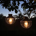 Zbliżenie na dwie świecące, wiszące lampy solarne w ogrodzie o zmierzchu. Lampy posiadają czarne daszki i zaokrąglone, druciane klatki chroniące żarówki imitujące dawne włókna węglowe. Ciepłe, złociste światło żarówek mocno kontrastuje z ciemnymi sylwetkami liści i gałęzi drzewa na pierwszym planie oraz z gasnącym, niebieskawo-szarym niebem w tle.
