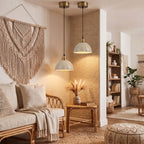 Jasny, przytulny salon urządzony w stylu boho. Z sufitu zwisają dwie kremowe lampy z karbowanymi kloszami i mosiężnymi detalami. Oświetlają one strefę wypoczynkową z rattanową kanapą z jasnymi poduszkami oraz mały, bambusowy stolik z wazonem pełnym suszonych traw. Na ścianie wisi duża, misternie pleciona makrama. W tle widać drewniany regał z dekoracjami i roślinami doniczkowymi.