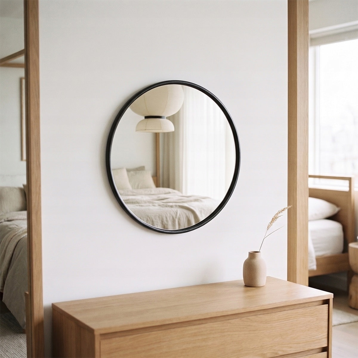Spokojna, minimalistyczna sypialnia w stylu japandi lub modern scandi. Na środku białej ściany wisi okrągłe lustro w czarnej oprawie, które odbija zarys łóżka z pościelą i charakterystyczną, papierową lampę sufitową. Pod lustrem znajduje się niska komoda wykonana z jasnego drewna dębowego, na której stoi jedynie mały, ceramiczny wazonik z pojedynczą, suszoną gałązką.