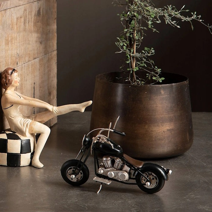 Czarno-srebrny model motocykla wykonany z metalu, postawiony na ciemnej posadzce. Z lewej strony znajduje się dekoracyjna figurka kobiety w stylu retro, siedzącej na czarno-białej pufie i zakładającej pończochę. W tle stoi duża, przecierana mosiężna donica, w której rośnie delikatna, pnąca roślina. Z lewej strony krawędzi kadru widać fragment drewnianej skrzyni.