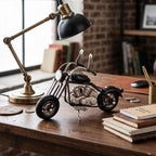 Metalowy model motocykla typu chopper stojący na drewnianym, klasycznym biurku. Czarno-srebrna figurka z detalami silnika i wysoką kierownicą jest głównym elementem kompozycji. Obok stoi mosiężna lampa gabinetowa z czarnym kloszem, notesy, złoty długopis w metalowym kubku oraz luźne monety. W tle widać ceglaną ścianę, okno oraz regał z książkami.