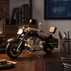 Stylowa aranżacja na ciemnym, drewnianym biurku w klasycznym gabinecie. Na pierwszym planie stoi metalowy, czarno-srebrny model motocykla typu chopper w stylu vintage. Obok niego znajduje się siatkowy przybornik z ołówkami i długopisami oraz skórzana podkładka. W tle, na regale, stoją rzędy oprawionych w skórę książek, a na ścianie wisi rozmyty, niebieski plakat (blueprint).