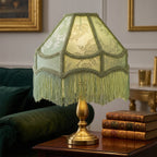 Włączona lampa stołowa z jasnomiętowym, żakardowym abażurem z frędzlami, zamocowanym na eleganckiej, mosiężnej podstawie. Lampa stoi na ciemnodrewnianym stoliku tuż obok stosu trzech oprawionych w skórę, starych książek. W rozmytym tle widoczny jest fragment ciemnozielonej, welurowej sofy oraz ściana ze sztukaterią i obrazami w złotych ramkach.
