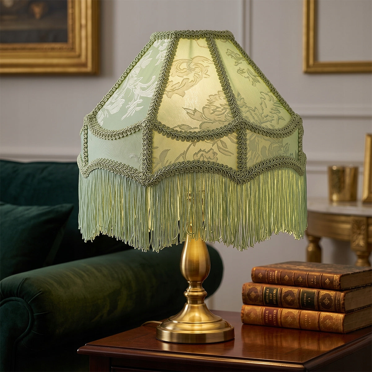 Włączona lampa stołowa z jasnomiętowym, żakardowym abażurem z frędzlami, zamocowanym na eleganckiej, mosiężnej podstawie. Lampa stoi na ciemnodrewnianym stoliku tuż obok stosu trzech oprawionych w skórę, starych książek. W rozmytym tle widoczny jest fragment ciemnozielonej, welurowej sofy oraz ściana ze sztukaterią i obrazami w złotych ramkach.