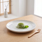 Czysta, minimalistyczna kompozycja w stylu azjatyckim. Na jasnym, ceramicznym talerzu z subtelnym nakrapianiem zaserwowano zielone ciastko matcha mochi, delikatnie oprószone herbacianym pudrem i ozdobione listkami świeżej mięty. Jasny, dębowy blat, drewniany widelczyk oraz rozmyte tło z japońskim akcentem nadają zdjęciu spokojny, harmonijny charakter w duchu zen.