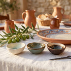 Zestaw trzech ceramicznych miseczek w apetycznej, śródziemnomorskiej aranżacji. Ustawione na jasnym, lnianym obrusie obok gałązki świeżych oliwek, doskonale sprawdzą się do serwowania oliwy, octu balsamicznego lub małych przekąsek typu tapas. W tle widać gliniane dzbanki, wzorzyste talerze oraz koszyk z pieczywem, co przywodzi na myśl leniwe, włoskie popołudnie (dolce vita).