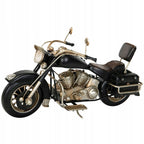 Zdjęcie produktowe na białym, gładkim tle (packshot). Dekoracyjny, metalowy model klasycznego motocykla (chopper) w kolorze czarnym z widocznymi srebrnymi i miedzianymi elementami silnika oraz układu wydechowego. Model ma szczegółowo oddane detale, takie jak skórzane siedzenie, oparcie dla pasażera, boczne sakwy, duże okrągłe przednie światło i lusterka. Ustawiony jest na wysuwanej nóżce.