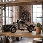 Metalowa figurka motocykla w stylu chopper stojąca na surowej, drewnianej półce wspartej na industrialnych rurach hydraulicznych. Z lewej strony motocykla znajduje się stary, czarny aparat fotograficzny w stylu retro (dalmierzowiec), a z prawej mały kaktus w glinianej doniczce. W tle widać rozmytą, ceglaną ścianę, duże okna ze szprosami oraz skórzaną kurtkę motocyklową wiszącą w oddali.