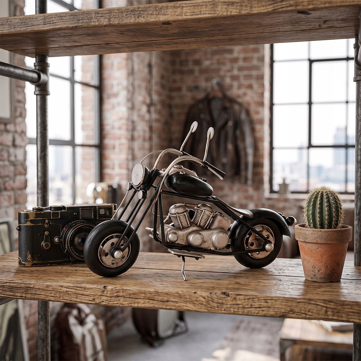 Metalowa figurka motocykla w stylu chopper stojąca na surowej, drewnianej półce wspartej na industrialnych rurach hydraulicznych. Z lewej strony motocykla znajduje się stary, czarny aparat fotograficzny w stylu retro (dalmierzowiec), a z prawej mały kaktus w glinianej doniczce. W tle widać rozmytą, ceglaną ścianę, duże okna ze szprosami oraz skórzaną kurtkę motocyklową wiszącą w oddali.
