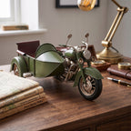 Dekoracyjna, metalowa figurka zielonego motocykla z wózkiem bocznym stojąca na drewnianym biurku. Obok modelu leży stos starych książek, eleganckie wieczne pióro, mosiężny kompas w drewnianym pudełku oraz klasyczna, mosiężna lampa gabinetowa. W tle widać jasne okno, nadające wnętrzu elegancki styl retro.