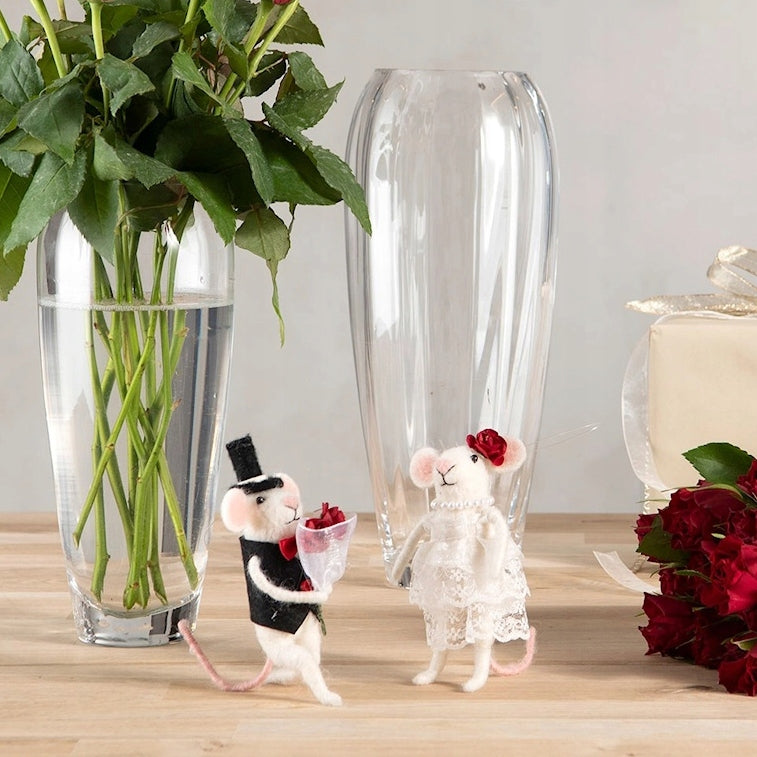 Romantyczna aranżacja na drewnianym stole. Filcowe figurki myszek w strojach ślubnych (pan młody we fraku, panna młoda w białej sukni) stoją obok wysokiego, szklanego wazonu, w którym umieszczono bukiet zielonych gałązek i czerwonych róż. Po prawej stronie widać fragment zapakowanego prezentu i kolejne czerwone róże.
