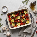 Zdjęcie z góry (flat lay) prezentujące pieczone warzywa w prostokątnej, ceramicznej formie do zapiekania o ciemnoszarych ściankach i jasnokremowym wnętrzu. Wewnątrz naczynia znajdują się plastry cukinii, pomidorki, czerwona papryka oraz czerwona cebula z gałązkami świeżego rozmarynu. Dookoła naczynia ułożono główkę czosnku, gałązki ziół, butelkę oliwy oraz miseczkę z solą morską