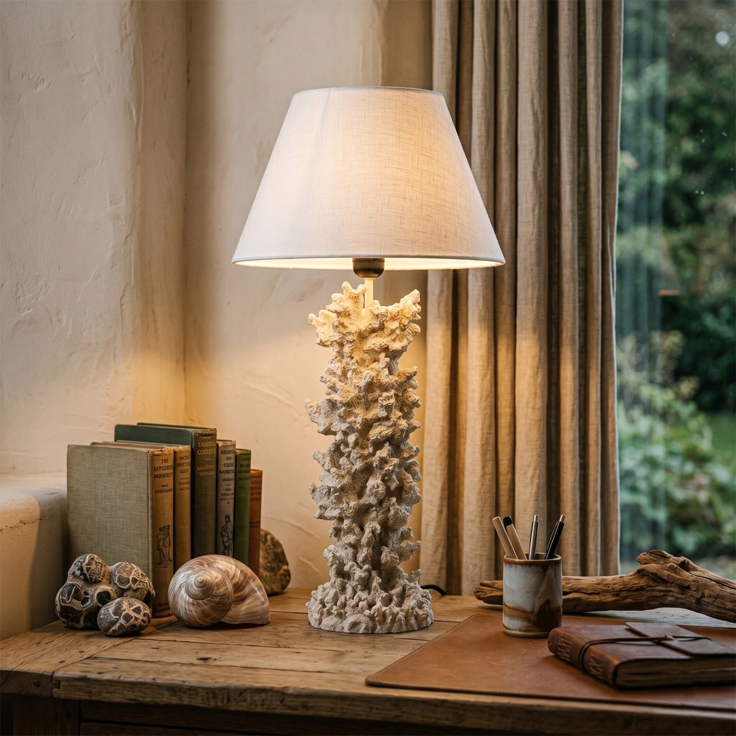 Lampa stołowa z koralowcową podstawą i jasnym abażurem, dająca ciepłe, nastrojowe światło. Naczynie stoi na starym, drewnianym biurku w otoczeniu przedmiotów nawiązujących do podróży i natury: starych książek, dużej muszli, kamieni, kawałka drewna wyrzuconego przez morze oraz skórzanego notesu. W tle widać okno z zasłoną, co nadaje scenie przytulny, gabinetowy charakter