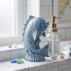 Ceramiczna figurka w kształcie ryby wyskakującej z fal, pokryta błyszczącym, niebieskim szkliwem. Ozdoba stoi na jasnym, marmurowym blacie w łazience. Obok niej znajduje się szklany dozownik na mydło z mosiężną pompką ozdobiony jutowym sznurkiem, a także ułożone w kostkę białe ręczniki, muszelki i kawałki kolorowego, morskiego szkła. W tle widać jasne okno, co dodaje aranżacji świeżości