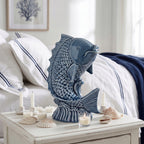 Błękitna, ceramiczna ryba wyeksponowana na białym, drewnianym stoliku nocnym w sypialni. Wokół figurki zaaranżowano kilka małych, białych świec, tealight w szkle oraz rozrzucone, naturalne muszle. W tle widać fragment łóżka z białą pościelą i narzutą w granatowe pasy, co doskonale współgra z marynistycznym charakterem (stylem coastal) całej kompozycji.