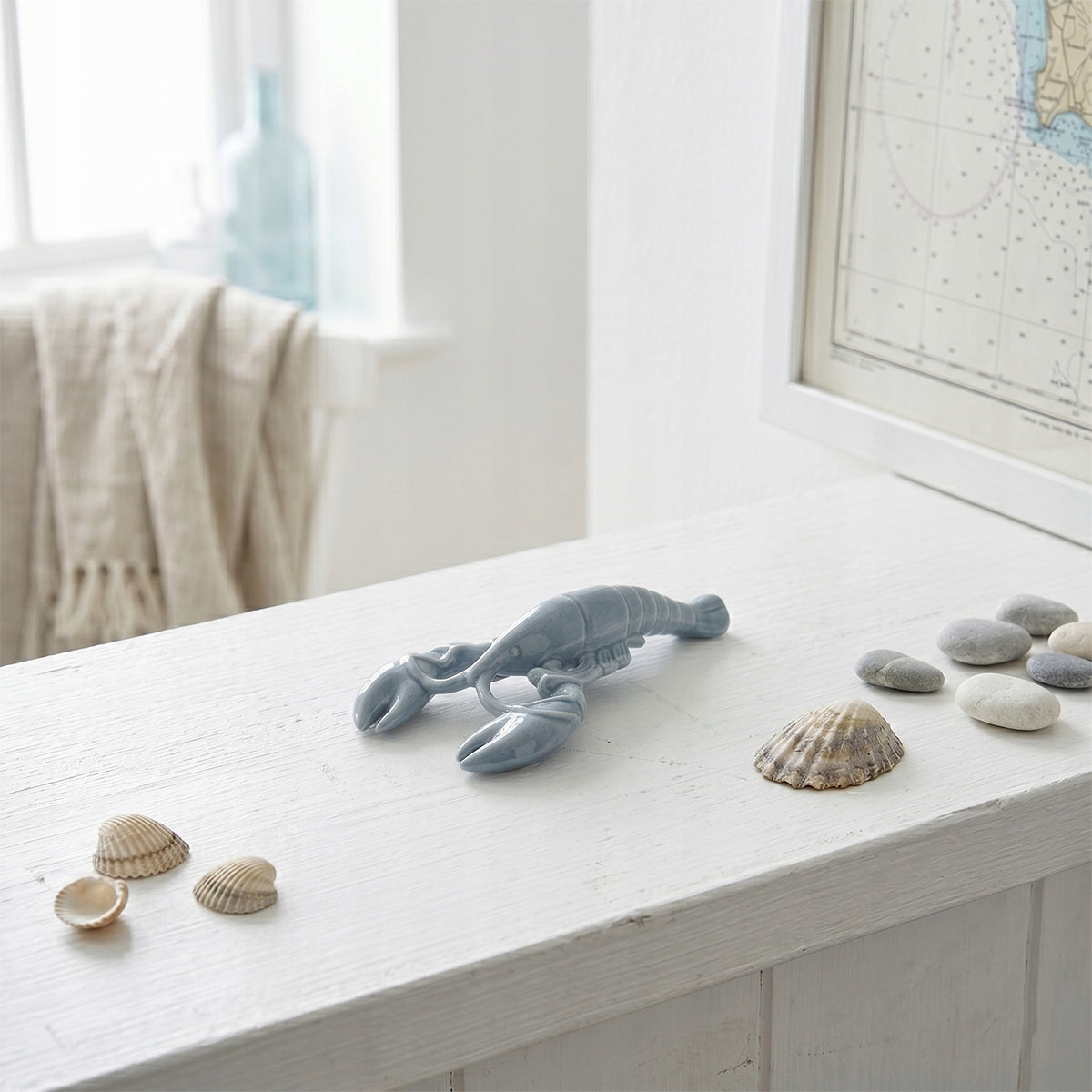 Ceramiczna figurka w kształcie niebieskiego homara wyeksponowana na białej, drewnianej komodzie. Obok ułożono drobne, gładkie kamienie (otoczaki) oraz muszle. W tle na ścianie wisi oprawiona w ramkę mapa żeglarska, a w głębi widać rozmyte okno i niebieską szklaną butelkę, co tworzy spójny, wakacyjny klimat aranżacji